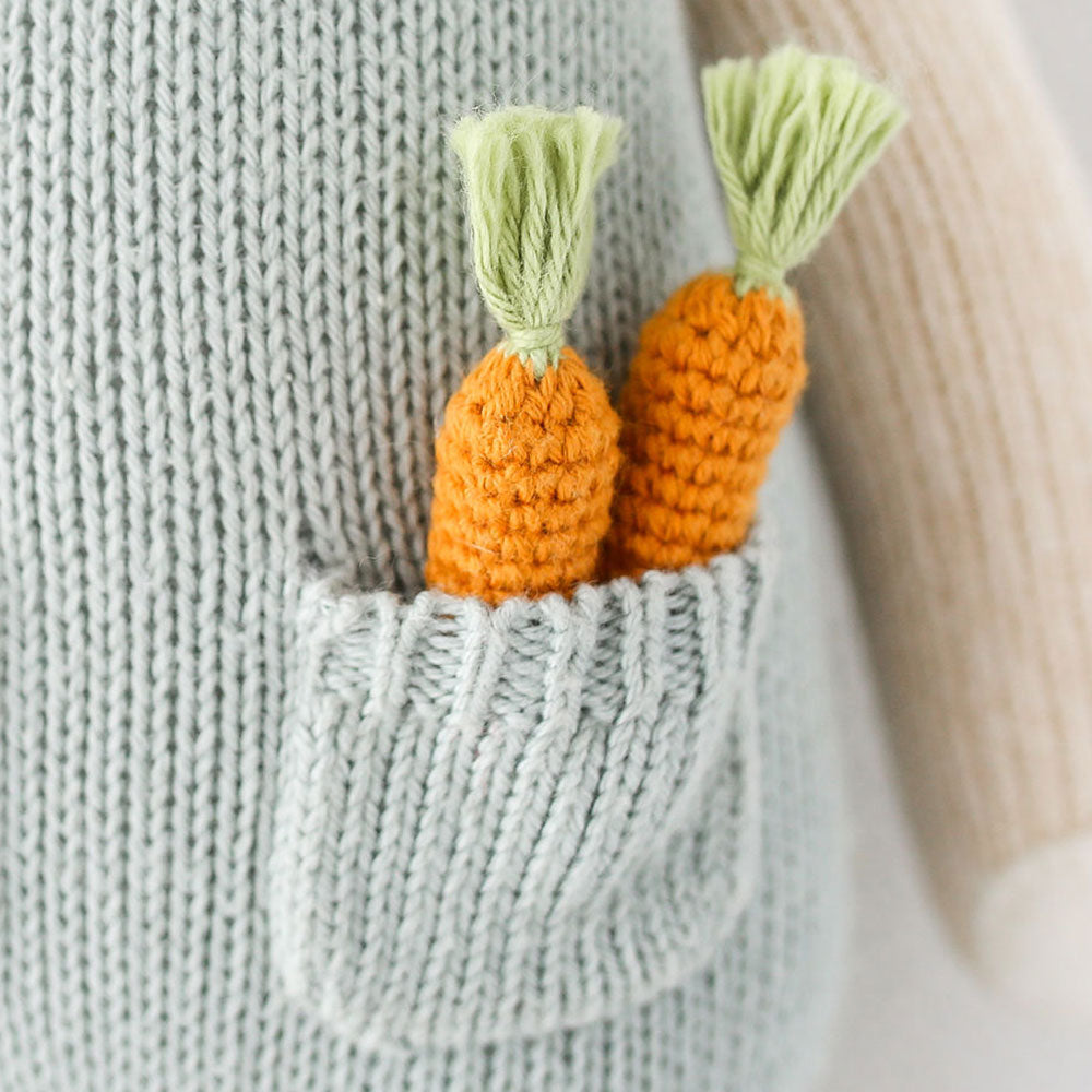 A close-up showing two hand-knitted carrots in the pocket of Henry’s overalls.