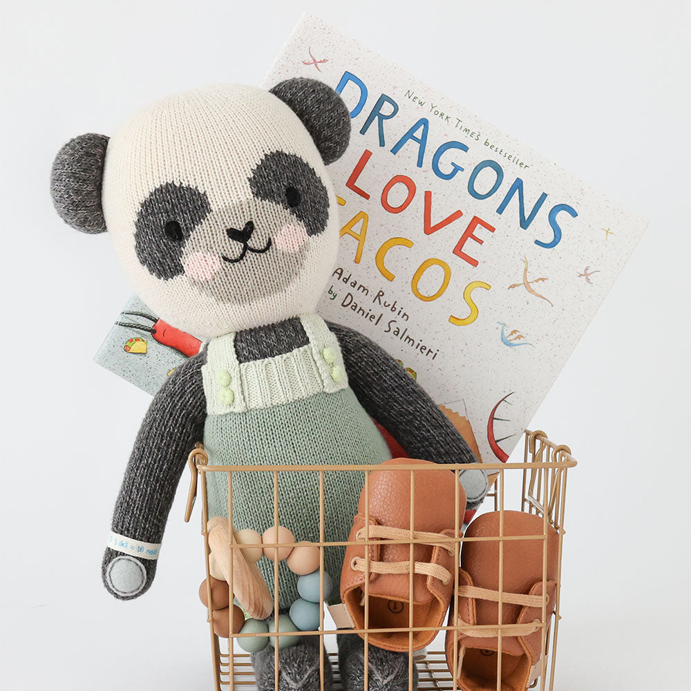 Paxton the panda sitting in a wire basket, alongside a children’s book, a pair of booties and a wooden infant’s toy.