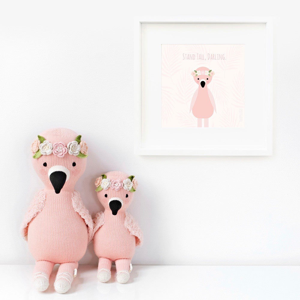 Two Penelope the flamingo stuffed dolls sitting beside a framed print with a picture of Penelope that says “Stand tall, darling.”